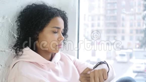 美国黑人妇女使用智能手表浏览坐在窗口视频的预览图
