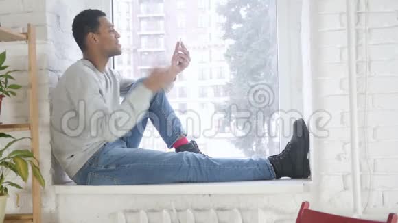 坐在窗边的非洲男人在智能手机上视频聊天视频的预览图