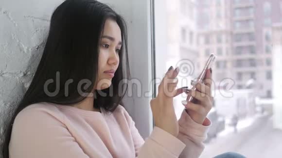 年轻的亚洲女性在浏览智能手机坐在窗边视频的预览图