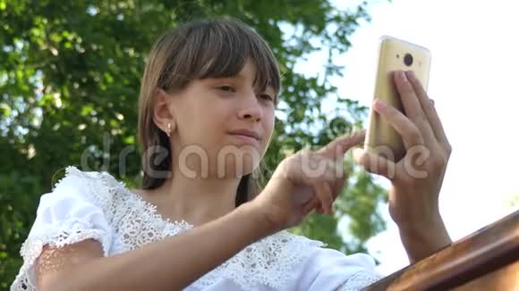 少女在长凳上用智能手机浏览照片在线查看电子邮件漂亮的女孩在智能手机上写着视频的预览图