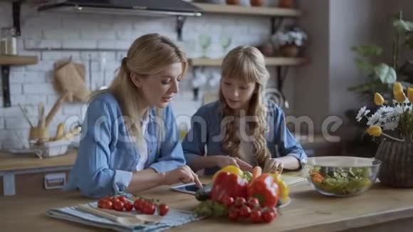 妈妈和女儿在明亮的大厨房里一起做饭女孩帮妈妈切菜妈妈看了菜谱视频的预览图