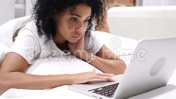 头痛沮丧的非裔美国妇女在床上的笔记本电脑上工作视频的预览图