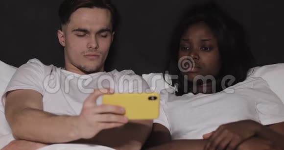多民族夫妇晚上一起在黄色智能手机上看视频躺在床上视频的预览图