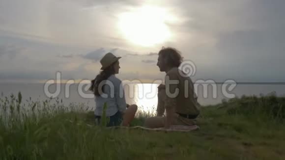 浪漫的假期有趣的笑声男人和女人交流坐在海边的绿草上天空的日落上有闪亮的水视频的预览图