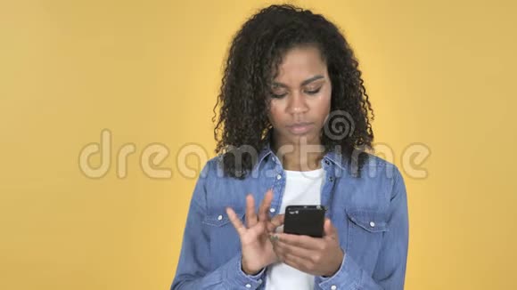 在黄色背景下使用智能手机的非洲女孩为成功而兴奋视频的预览图