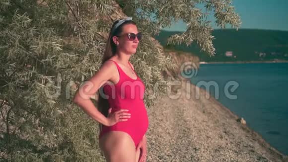 怀孕的女孩穿着红色的一件泳衣和眼镜靠近一棵树站在海滩上微笑着日落视频的预览图