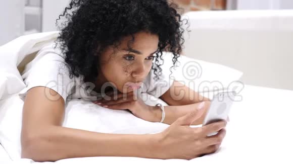 疲惫的非洲裔美国妇女躺在床上滚动和浏览智能手机视频的预览图
