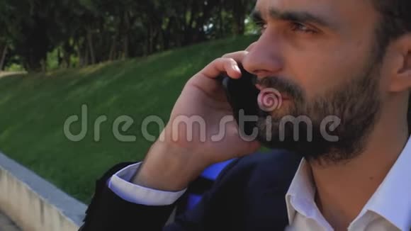 一个有魅力的商人坐在公园的长椅上留着胡子等待电话长胡子的男人视频的预览图