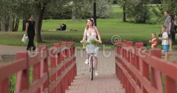 穿着白色夏装的浪漫性感女孩骑着自行车在森林公园的木桥上散步视频的预览图