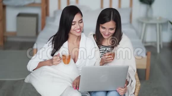 两个年轻的女姐妹在家里用笔记本电脑看喜剧电影喝茶视频的预览图
