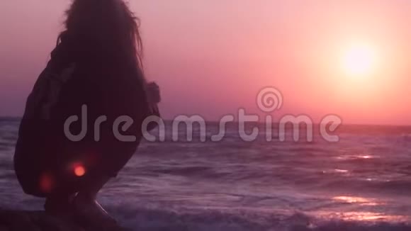 美丽的女孩坐在石头上看着粉红色的日落看着深蓝色的大海视频的预览图