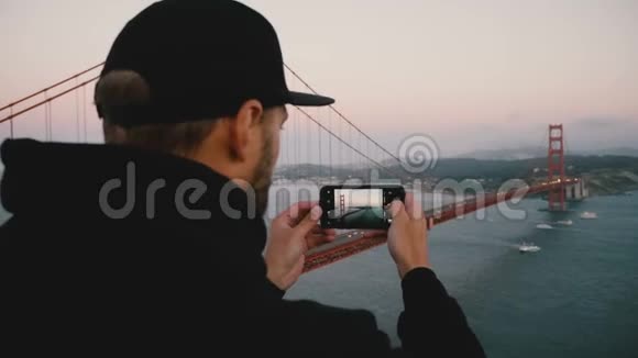 后景年轻留胡子的男人穿着黑色连帽衫和帽子拍摄智能手机照片日落金门大桥旧金山视频的预览图