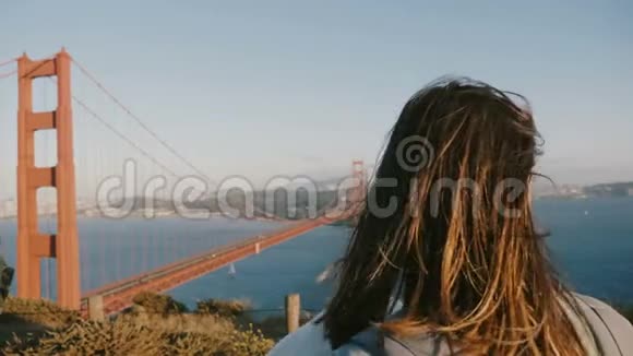 在金门大桥欣赏史诗般的日落时一位头发随风飘动的年轻旅游妇女的特写后景镜头视频的预览图