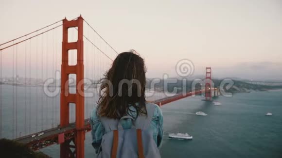 后景年轻快乐的和平旅游妇女背包和风在她的头发观看标志性的金门大桥景观视频的预览图