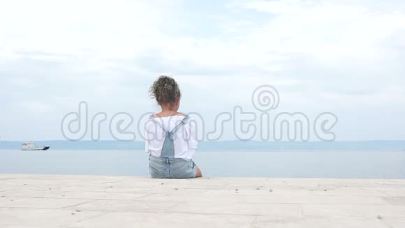 女孩独自坐在海湾的岸边看着船离开一个卷发男孩进来坐在他旁边视频的预览图