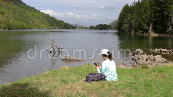 年轻的旅游女孩坐在湖边用手机拍照视频的预览图
