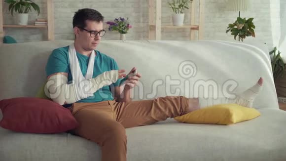 一个手臂和腿骨折的年轻人坐在沙发上用智能手机的画像视频的预览图