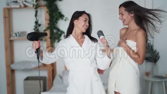两位年轻女士穿着白色浴袍跳舞带吹风机唱歌吹干头发开怀大笑视频的预览图