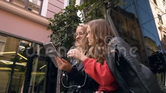 夏天两个漂亮的白人年轻女孩坐在玻璃大厦附近看着平板电脑上的东西视频的预览图