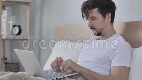 快乐男人在床上使用笔记本电脑庆祝成功视频的预览图