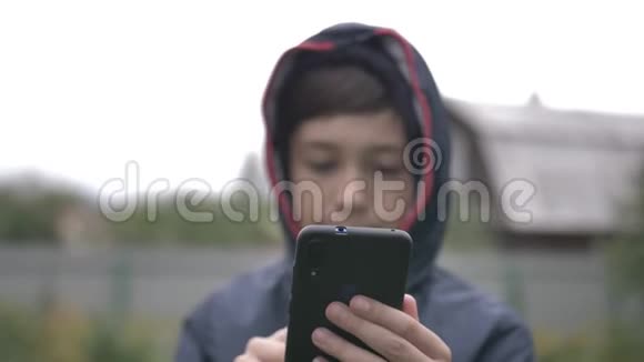 风帽里的滑稽男孩在户外用手机关门秋天多云天气移动摄像机视频的预览图