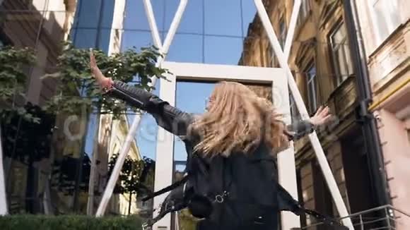 开朗的白种人女孩留着长发戴着时髦的眼镜高兴地走在玻璃大厦附近的城市街道上视频的预览图