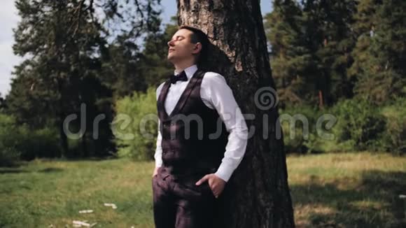 时髦的帅哥站在大自然中背靠着一棵树闭上眼睛阳光照在他的脸上视频的预览图