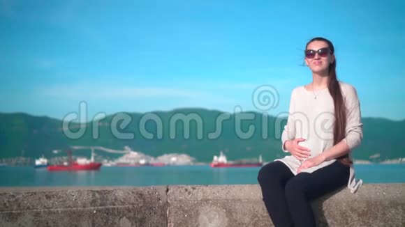 夏天天气晴朗年轻的怀孕迷人的女孩戴着太阳镜女孩坐在靠近大海的堤岸上视频的预览图