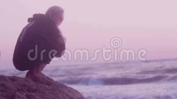 日出时美丽的女孩坐在石头上看着相机海波浪背景上的白色天空视频的预览图