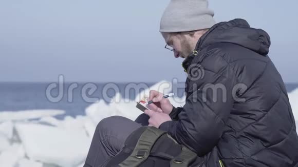 英俊的金发胡须男坐在冰间把他的观察写在笔记本上冰川上的极地探险家视频的预览图