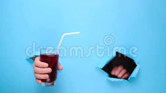 雌性手拿着一杯新鲜的红色果汁用吸管穿过撕裂的蓝色纸墙果饮料和健康营养视频的预览图