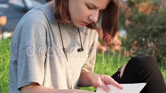 穿着灰色T恤的小女孩坐在公园的草地上环顾四周看一本带有讲稿的笔记本视频的预览图