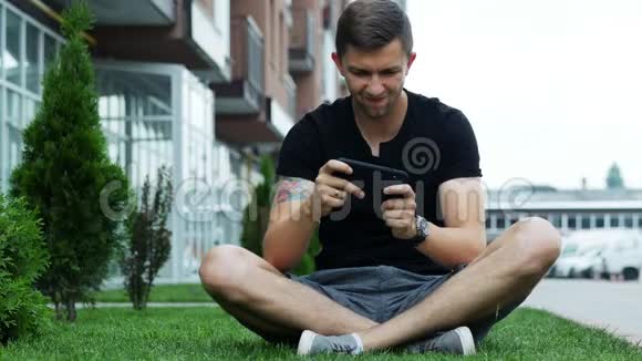 快乐男人在智能手机上玩手机游戏坐在房子附近的草地上视频的预览图