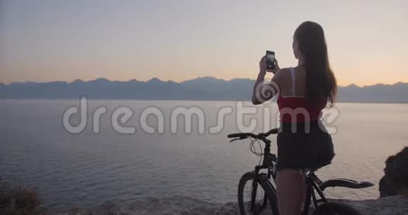 美丽的女士骑着自行车在悬崖上用智能手机在海上拍摄日落视频的预览图