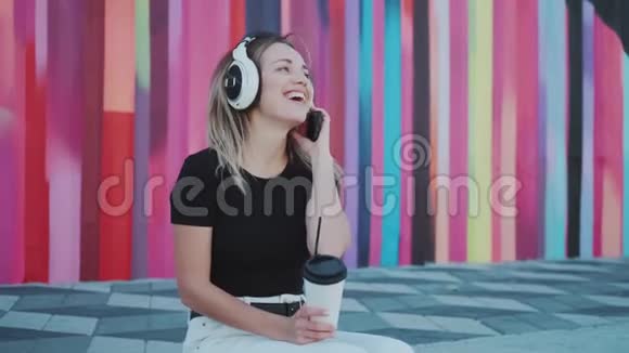 快乐无忧无虑的女孩在户外对着彩色的墙壁戴着大耳机听音乐视频的预览图