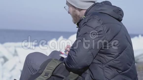 英俊的金发胡须男坐在冰间把他的观察写在笔记本上冰川极地探险家视频的预览图