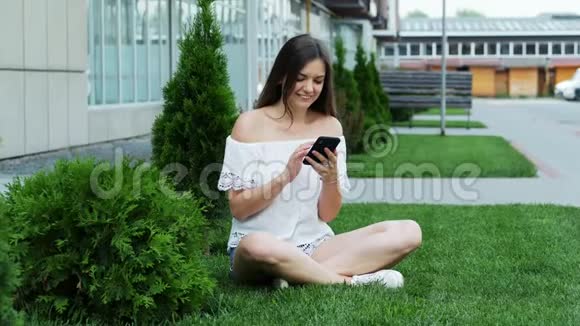 年轻快乐的女人坐在酒店附近的草坪上用电话留言视频的预览图