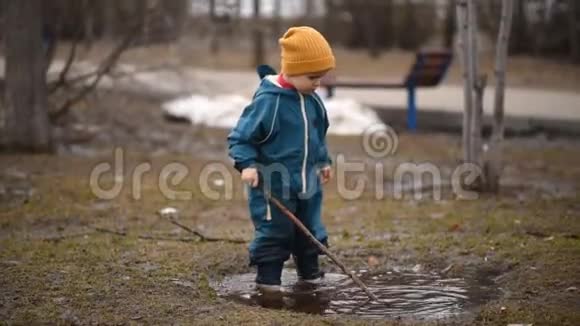 快乐的孩子穿着防水服手里拿着棍子在水坑里跳视频的预览图