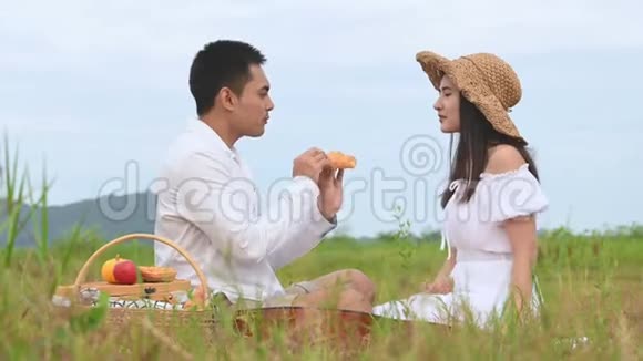 亚洲年轻夫妇在绿色草地野外背景下野餐男人把苹果给女人在调情蜜月之间吃饭视频的预览图