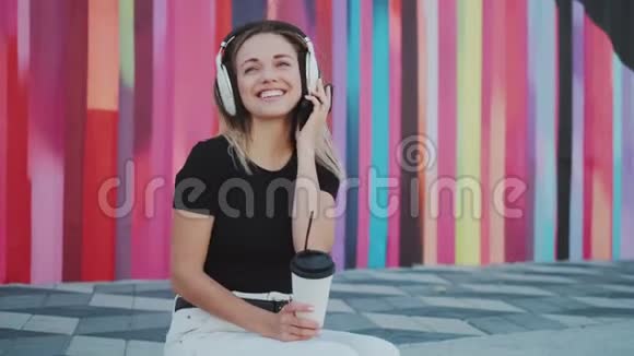 一位迷人的女孩带着耳机听音乐和在户外喝咖啡的肖像视频的预览图