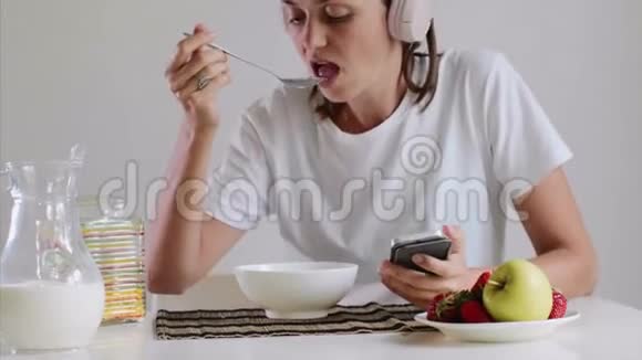 女人吃麦片用耳机听音乐浏览智能手机视频的预览图