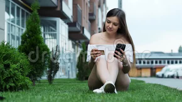 美丽的年轻女子通过电话进行网上购物并在附近的草地上输入信用卡号码视频的预览图