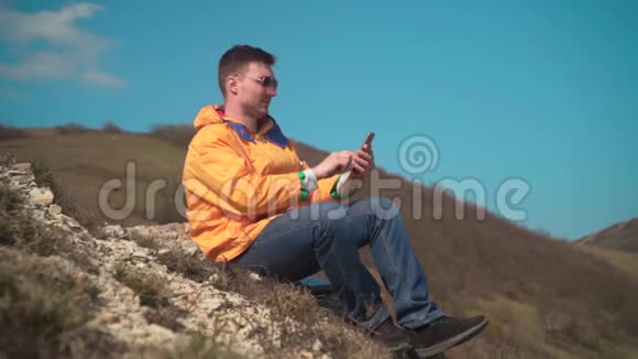 一个穿着黄色夹克蓝色牛仔裤和眼镜的男人坐在山上欣赏风景在电话上拍摄视频视频的预览图