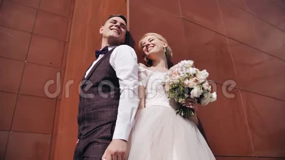 一对漂亮的年轻夫妇站在一座高楼的棕色墙附近恋人们微笑视频的预览图