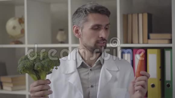 一幅穿着白色长袍的英俊男子在镜头中提出胡萝卜和西兰花的肖像现代熟练的医生视频的预览图
