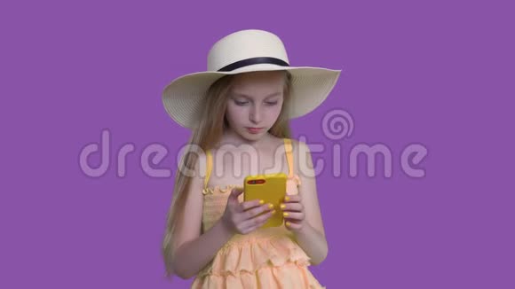 戴着夏季帽子的金发女孩正在紫色墙背景上浏览智能手机视频的预览图