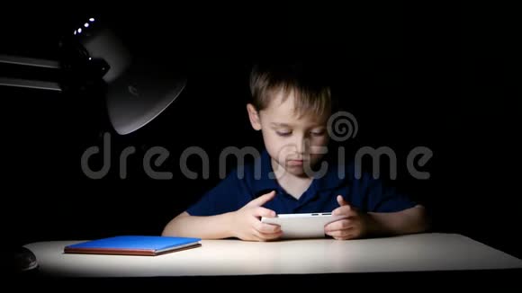 一个孩子晚上在家看卡通片他用智能手机坐在桌子旁被一盏灯照亮视频的预览图