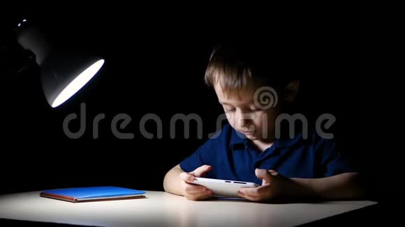 一个孩子晚上在家里开心地用智能手机看着一幅漫画坐在一张灯点亮的桌子旁微笑着视频的预览图