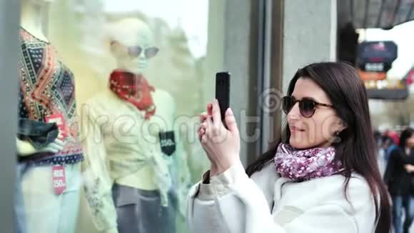 时尚的购物者女士用智能手机在商店时尚服装橱窗中拍照视频的预览图