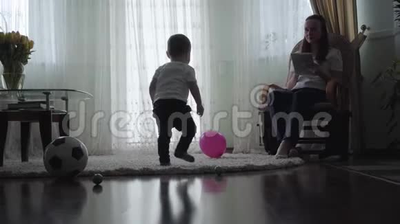 年轻的母亲坐在家里的沙发上旁边的女人是一个小男孩在地板上玩一个大球视频的预览图